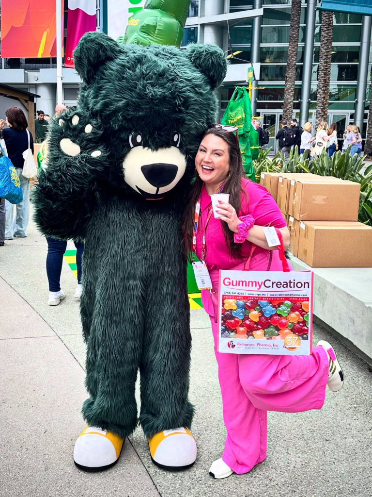 Tiffany from Your Neighborhood Vegan posing with the Grüns gummy bear mascot outside the Anaheim Convention Center at Natural Products Expo West 2026.
