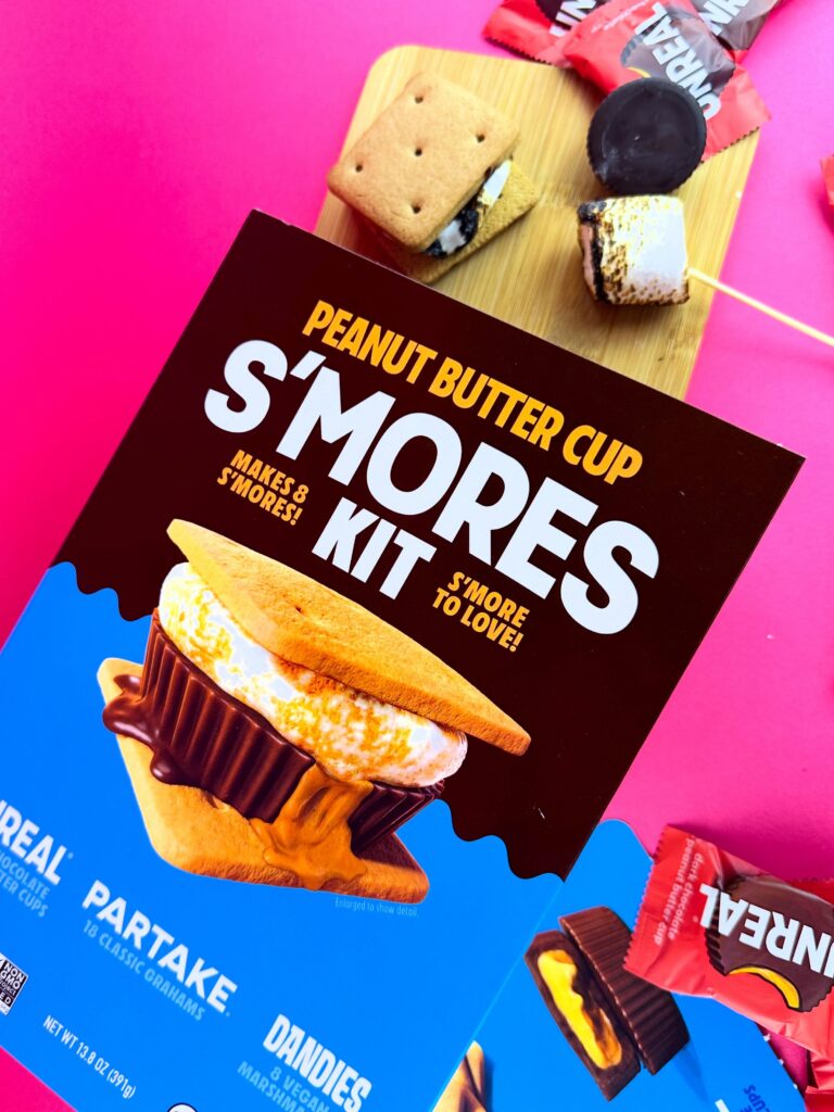 Vegan peanut butter cup s’mores kit box styled with toasted marshmallows, graham crackers, and vegan peanut butter cups on a vibrant pink background.