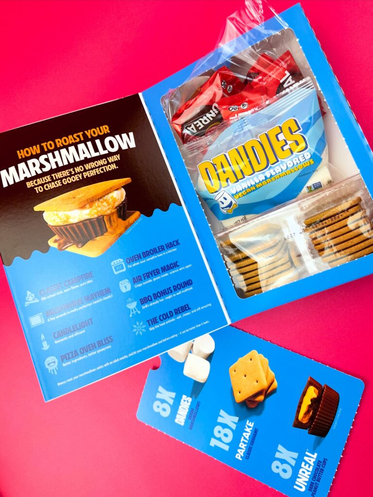 Open vegan s’mores kit box showing graham crackers, Dandies marshmallows, instructions, and vegan peanut butter cups on a bright pink background.