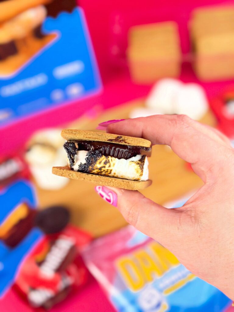 Hand holding a vegan peanut butter cup s’more made with melted Dandies marshmallows, graham crackers, and a chocolate peanut butter cup in front of bright pink background and s’mores kit ingredients.