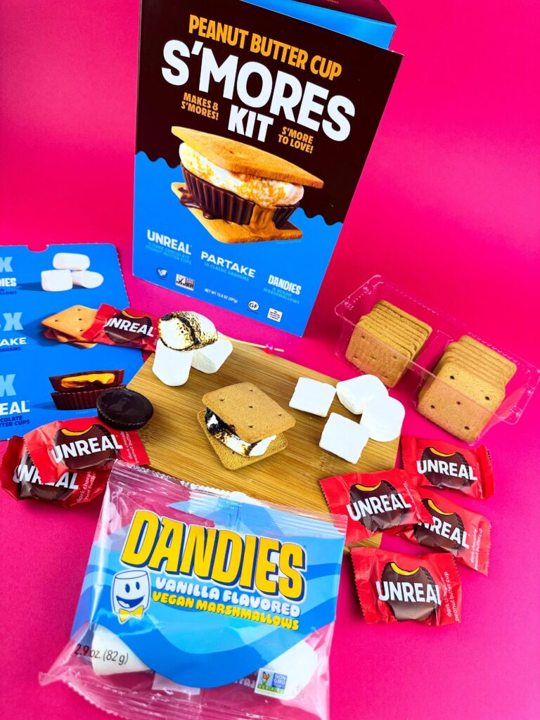 Flat lay of a vegan peanut butter cup s’mores kit with Dandies vegan marshmallows, graham crackers, Unreal peanut butter cups, and bright pink background.