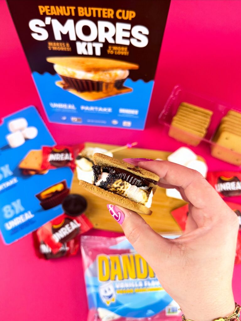 Hand holding a vegan peanut butter cup s’more in front of a bright pink flat lay featuring Dandies marshmallows and Unreal peanut butter cups.