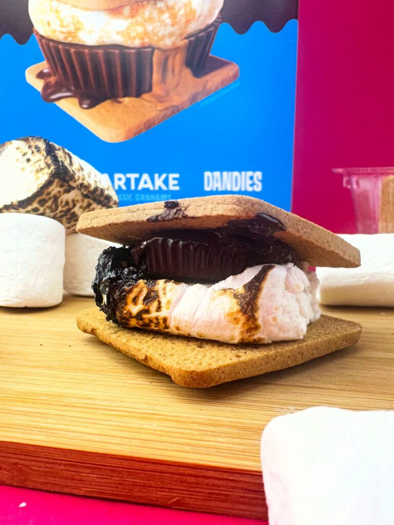 Close-up of a vegan peanut butter cup s’more with toasted marshmallow and melted chocolate on a graham cracker, styled with Dandies marshmallows and kit packaging in the background.