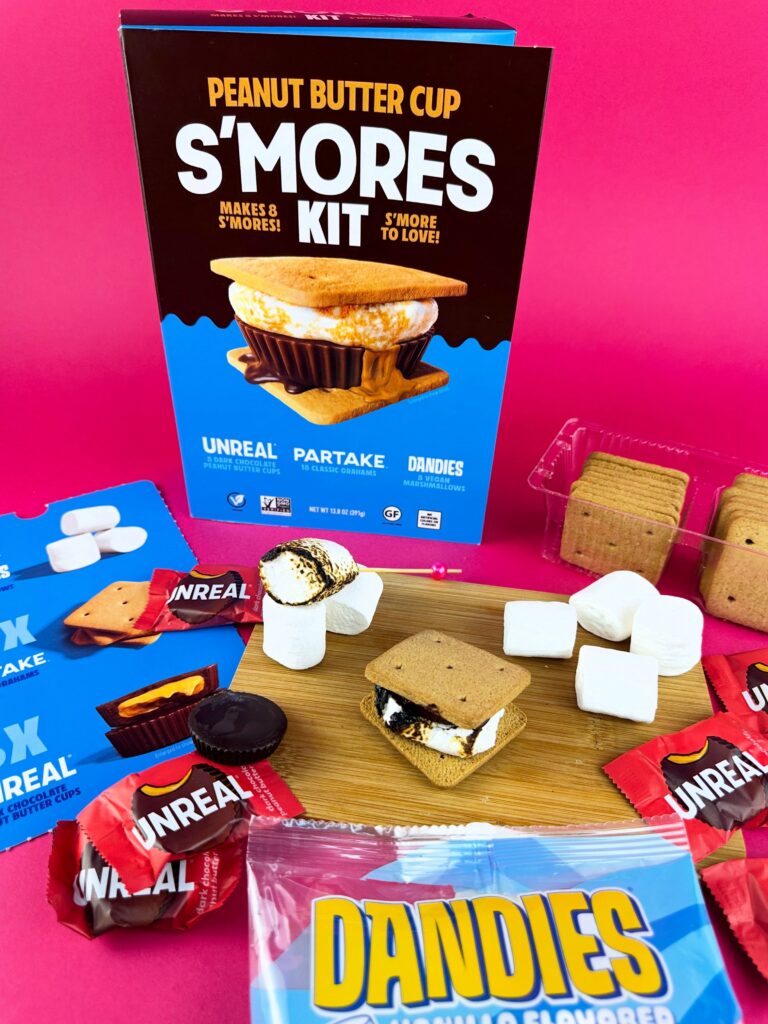 Vegan peanut butter cup s’mores kit packaging surrounded by marshmallows, graham crackers, vegan peanut butter cups, and pink background styling.