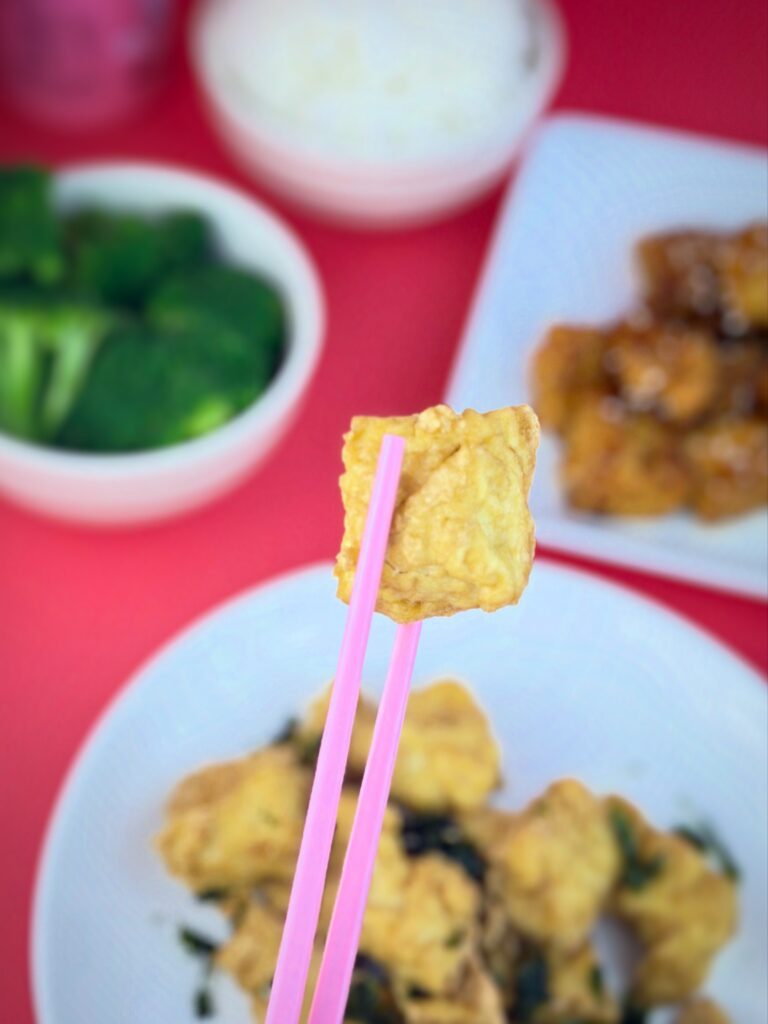 Piece of Blackbird Foods Original Crispy Tofu held with pink chopsticks with rice, broccoli, and tofu dishes blurred in the background.