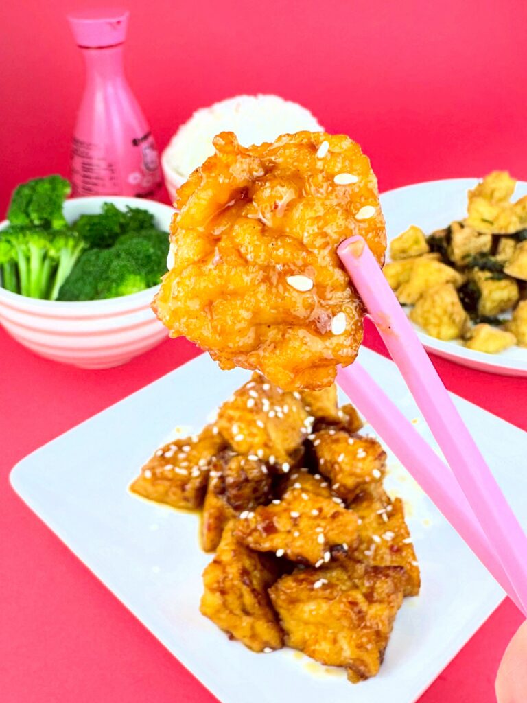 Piece of Blackbird Foods General Tso's Crispy Tofu held with pink chopsticks above a plate of sesame-topped tofu.