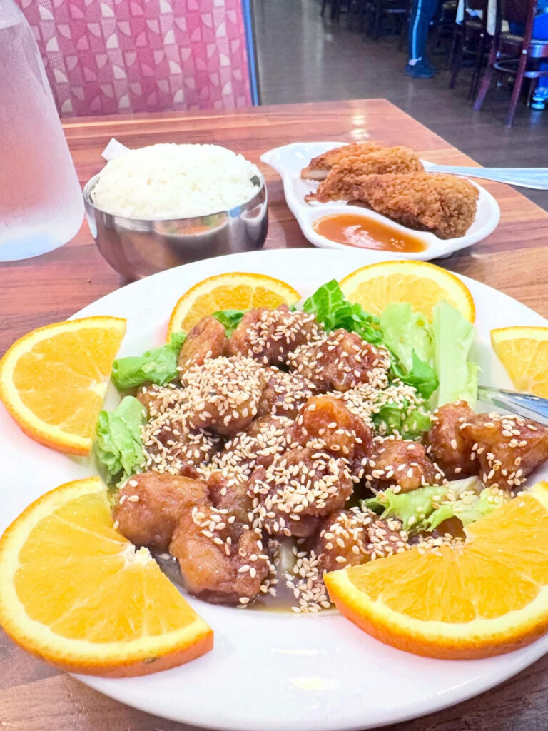 Sweet Orange entrée at Blossom Vegan featuring crispy soy protein coated in orange sauce, topped with sesame seeds and served with rice and fresh orange slices.