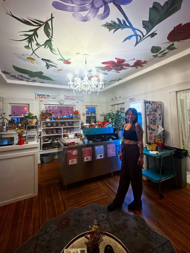 Interior of Alchemist Café & Elixir Bar featuring colorful ceiling artwork, bakery counter, dining space, and owner Candace.