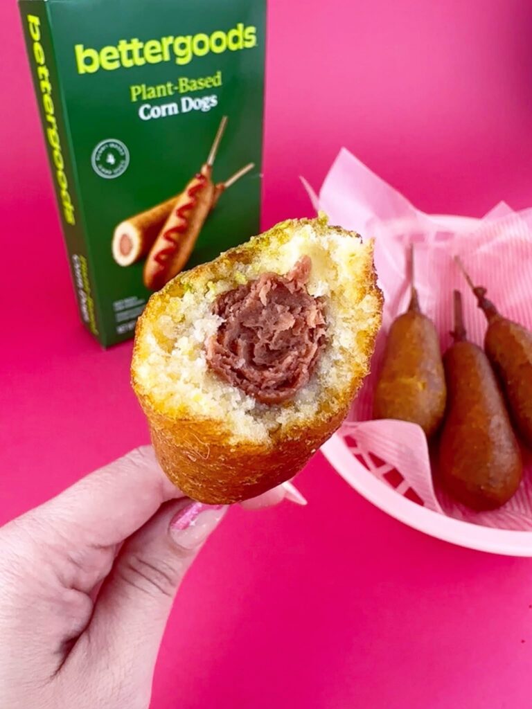 Close-up of a bitten Bettergoods Plant-Based Corn Dog showing the inside texture, with two cooked corn dogs and the Bettergoods box in the background.
