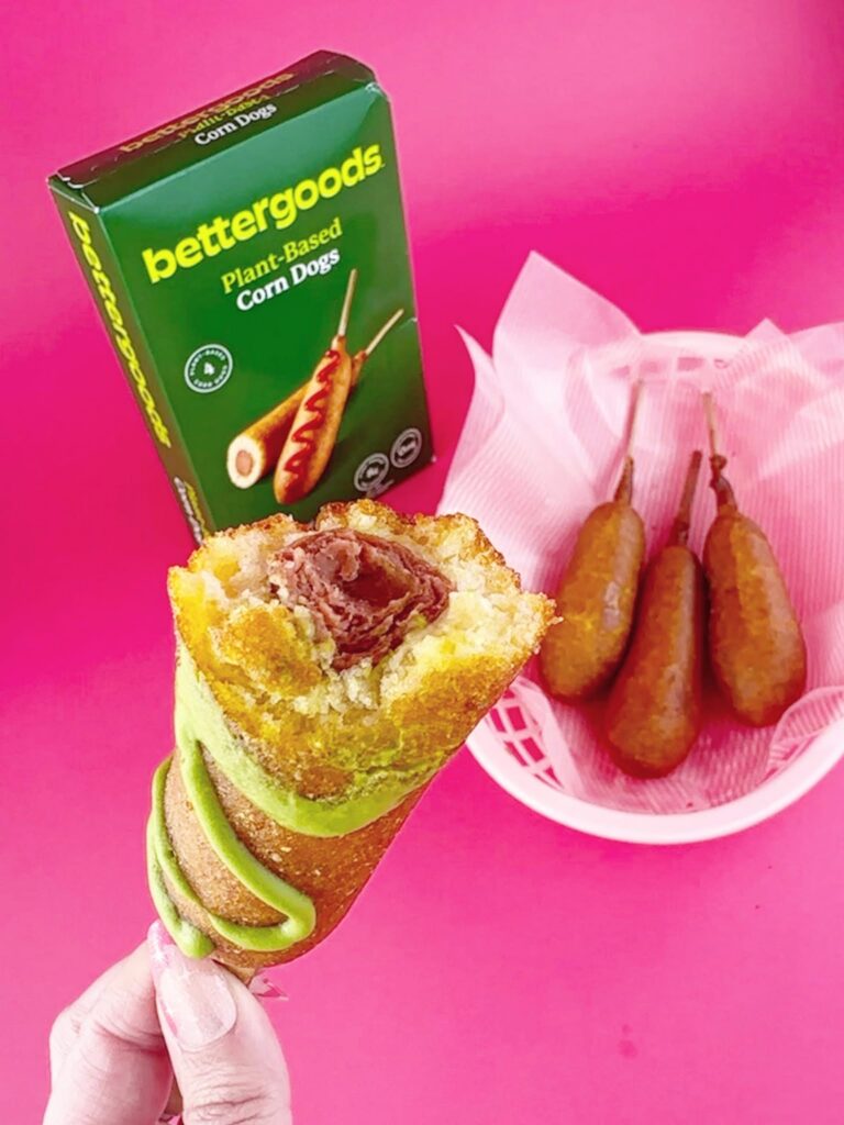 Cross-section of a Bettergoods Plant-Based Corn Dog showing the fluffy cornmeal coating and plant-based hot dog, with additional corn dogs and the product box behind it.