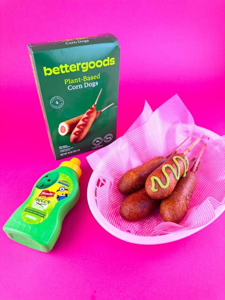 Bettergoods Plant-Based Corn Dog topped with green mustard beside two corn dogs in a pink basket, with the product box and French's Goomi Green Mustard bottle in the background.