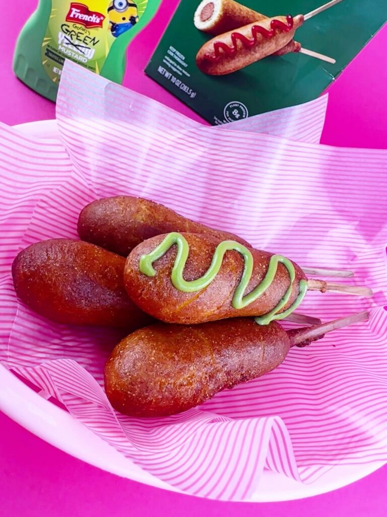 Three cooked Bettergoods Plant-Based Corn Dogs served in a pink paper-lined basket with the Bettergoods box displayed behind them.
