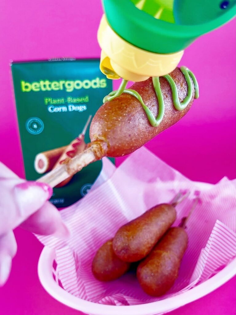 Bettergoods Plant-Based Corn Dog being drizzled with green mustard from a bottle above a pink basket of cooked corn dogs.