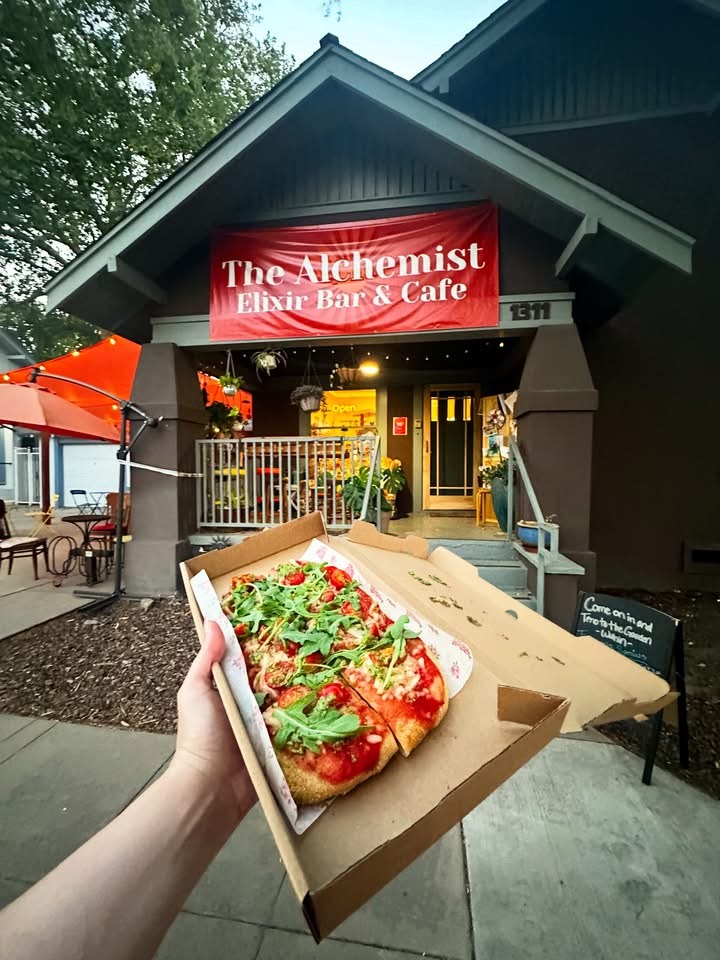 Hand holding a vegan Margarita Flatbread in front of Alchemist Café & Elixir Bar in downtown Modesto.