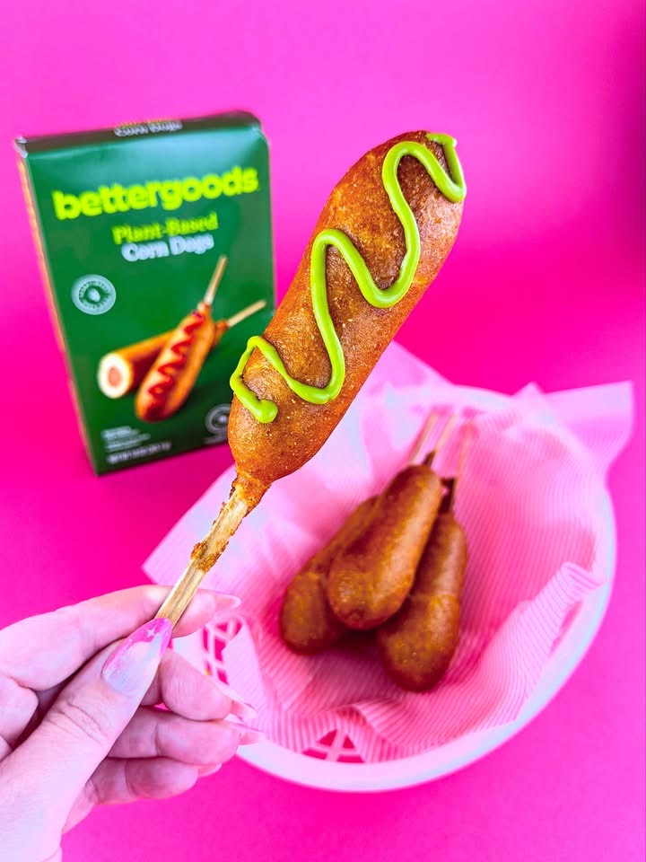 Bettergoods Plant-Based Corn Dog held over a pink basket with a zigzag of green mustard, with the Bettergoods box in the background.
