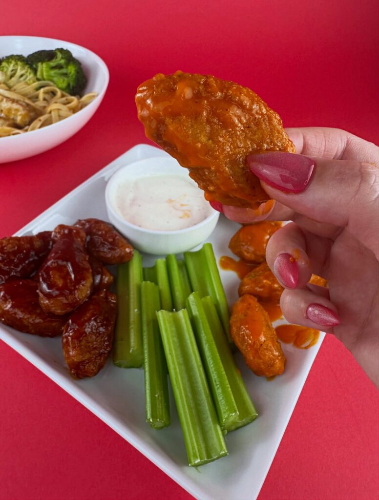 Hand holding a MorningStar Farms Buffalo Chik'n Wing dipped in vegan ranch over a platter of wings and celery.