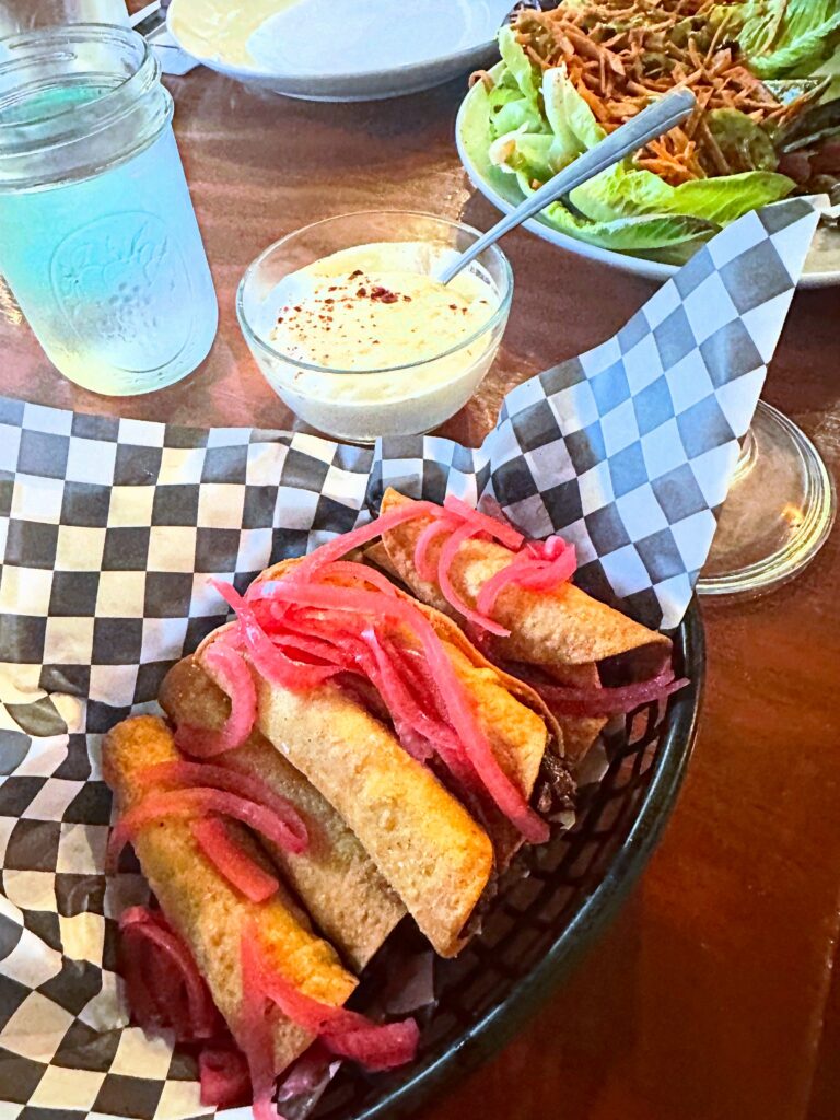 Jackfruit carnitas taquitos topped with bright pink pickled onions and served with vegan cheese sauce.