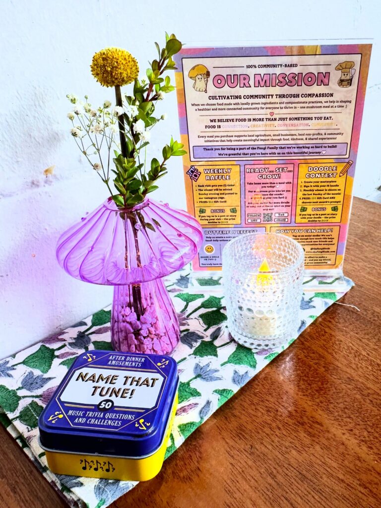 Fungi Foods table display featuring flowers, a community raffle sign and community conversation card game.
