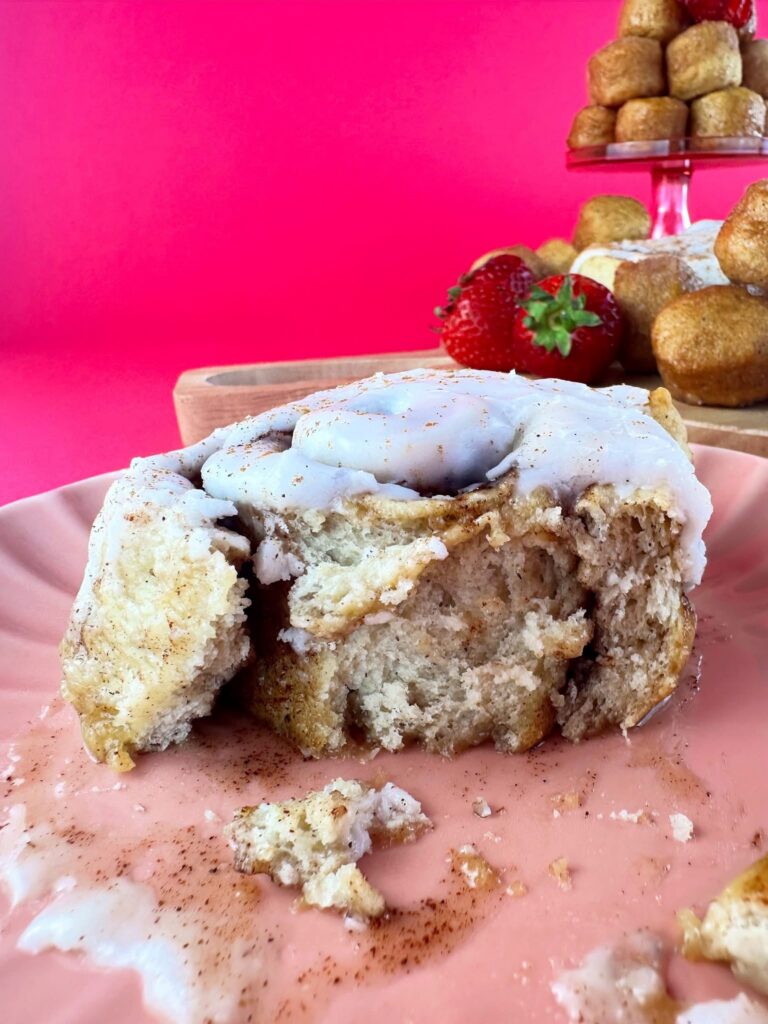 Partially eaten vegan gluten-free cinnamon roll showing the soft cinnamon-filled center with donut holes and strawberries behind it.