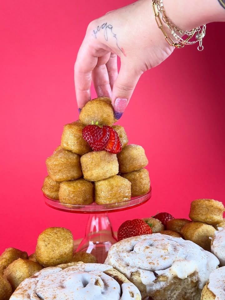Hand stacking vegan gluten-free glazed donut holes beside cinnamon rolls and strawberries from Gluten Gone Goodies.