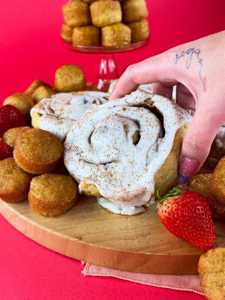 Hand holding a vegan gluten-free cinnamon roll over a platter of cinnamon rolls, donut holes and strawberries.