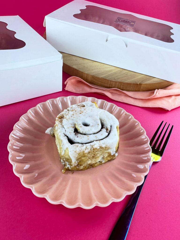 Vegan gluten-free cinnamon roll from Gluten Gone Goodies served on a pink plate beside its bakery box.