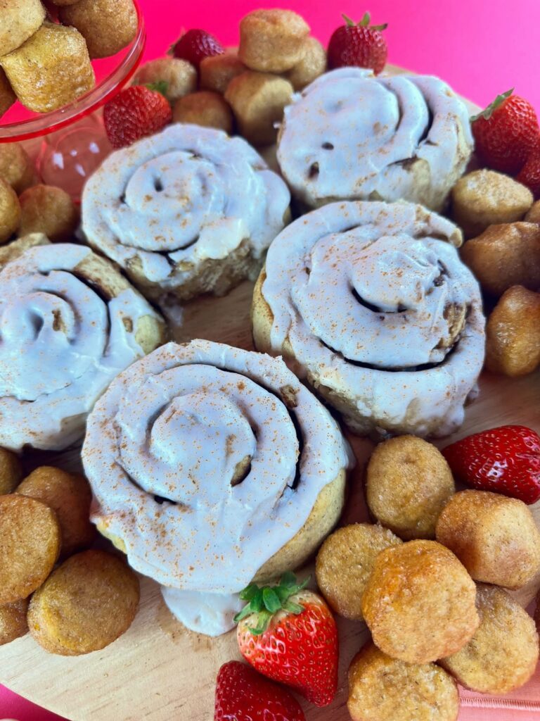 Close-up of iced vegan gluten-free cinnamon rolls surrounded by glazed donut holes and strawberries.