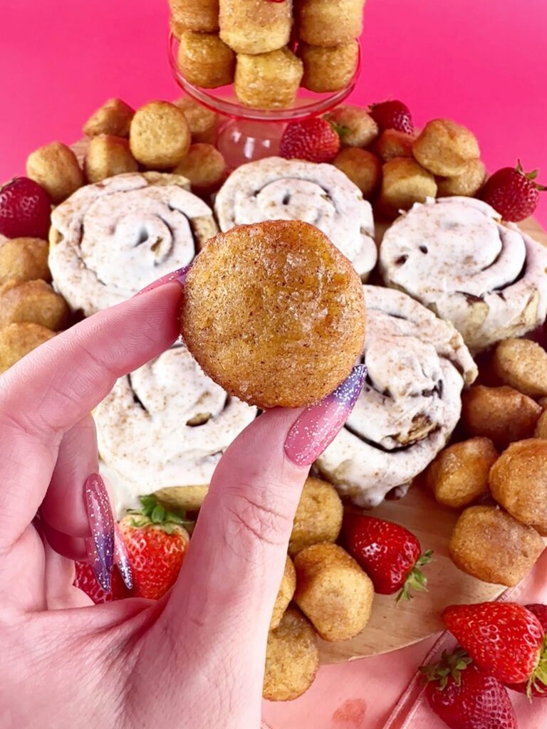 Vegan gluten-free cinnamon roll held over a platter of cinnamon rolls, glazed donut holes and strawberries.