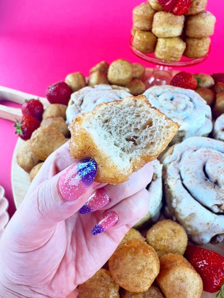 Hand holding a glazed vegan donut hole in front of Gluten Gone Goodies cinnamon rolls and donut holes.