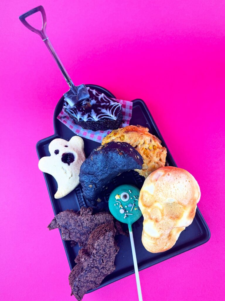 Assorted vegan Halloween treats from Vegan Dollhouse, including skull pizza, moon scone, ghost hand pie, bat brownie, graveyard cup, and cookie pop.